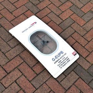 May include: A white airplane window clock with a black clock face and hands. The clock is labeled "G-EUPB AIRBUS A319-131 MSN 1115 Test Reg D-AVYT 27 October 1999 01 August 2020".