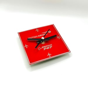 May include: Red square clock with black hands and silver accents. The clock face features the text "British Airways" and "Boeing 747".