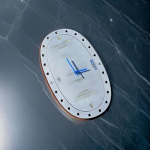 May include: Oval-shaped metal clock with a silver face and copper-colored trim. The clock features blue hands and the text "BRITISH AIRWAYS" and "BOEING 747-436". The clock also has the text "MAY 1996" and "NOVEMBER 2020".