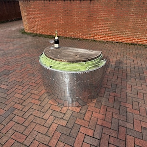 May include: A silver, round, metal table with a wooden top. The table is sitting on a brick patio. A bottle of champagne and a champagne glass are on the table.