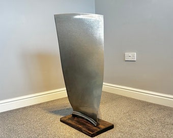 Puede incluir: Una gran hélice de metal plateado montada sobre una base de madera. La pala es curva y tiene una superficie lisa y pulida.