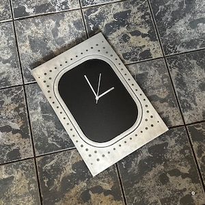 May include: A silver metal clock with a black face and white hands. The clock is shaped like an airplane window and has a textured surface.