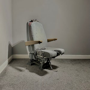 May include: A grey aircraft seat with a grey fabric covering and a wooden armrest. The seat has a metal frame and is in a room with grey carpet.