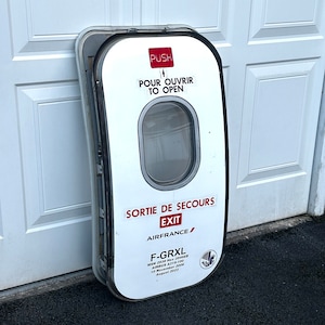 May include: White aircraft emergency exit door with a round window. The door has black trim and red and black text that reads "Sortie de Secours Exit Airfrance/ F-GRXL MSN 2938 Hex 294688 Airbus A319-100 10 November 2006 August 2023".