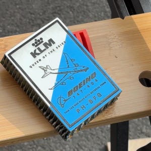 Può includere: Un'etichetta di aereo KLM blu e bianca con il testo "KLM Queen of the Skies" e "Boeing 747-404". L'etichetta include anche il testo "This tag was made with material from PH-BFG".