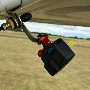 May include: A black GoPro camera is attached to an aircraft wing with a custom bracket. The bracket has silver, red, and brass-coloured parts. The camera is angled downwards, capturing the view below.