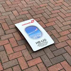 May include: A white sign with a red Swissair logo and a picture of an airplane in a window frame. The sign reads "HB-IJG AIRBUS A320-214 MSN 566 Hex 502CCA 22 November 1995 11 June 2021".