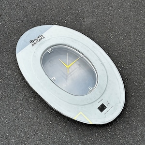 May include: A white circular clock with a gray face and black numbers. The clock is surrounded by a gray ring with the words "Royal Air Force" and "ZAMY" printed on it. The clock is on a gray surface.