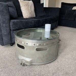 Könnte beinhalten: Ein runder, silberner Couchtisch aus einem wiederverwendeten Flugzeugteil. Der Tisch hat eine Glasplatte und steht auf einem grauen Teppich. Eine weiße Kerze mit dem Wort "Inspiration" darauf steht auf dem Tisch.