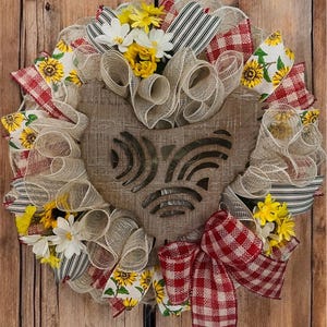 May include: A rustic farmhouse-style wreath with a wooden owl cutout at the center. The wreath features beige mesh, red and white gingham ribbons, sunflower and daisy accents, and black and white striped ribbons. The wreath is displayed against a wooden background.