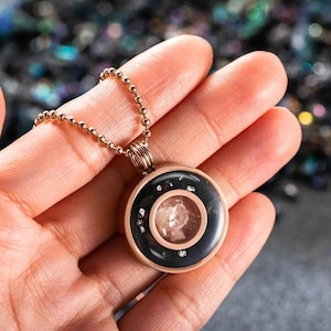May include: A rose gold chain necklace with a round pendant. The pendant is black with a clear center that contains small, white crystals.