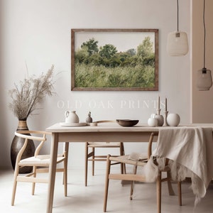 May include: A framed watercolour painting of a green field with trees and a house in the distance. The painting is hanging on a white wall above a wooden dining table with chairs. The table is set with a bowl, a pitcher, and other decorative items.
