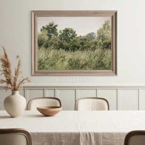 May include: A framed print of a landscape painting depicting a field of tall grass and trees in the background. The print is hanging on a white wall above a white table with a wooden bowl and chairs.