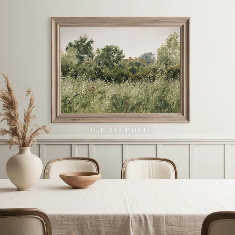 May include: A framed print of a landscape painting depicting a field of tall grass and trees in the background. The print is hanging on a white wall above a white table with a wooden bowl and chairs.