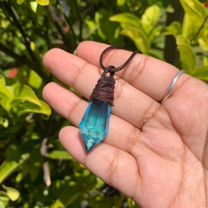May include: A blue crystal pendant necklace with a brown cord. The crystal is a faceted, elongated shape, wrapped with brown twine at the top. The background is blurred green foliage.