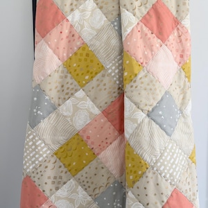 May include: A quilt made of various fabrics in shades of pink, yellow, and gray. The quilt is made in a diamond pattern and has a modern, geometric design.