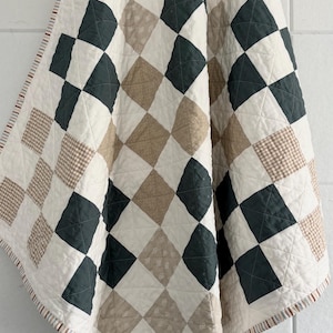May include: A quilted blanket with a diamond pattern in cream, dark green, and tan. The blanket has a striped border in red, brown, and cream. The quilt is made of soft fabric and is suitable for a bed or sofa.