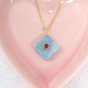 May include: A gold chain necklace with a light blue, four-petal pendant and a small red gemstone in the center. The necklace rests on a pink heart-shaped dish, set against a white background.