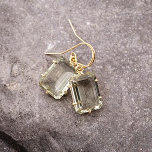 May include: A pair of gold-toned hook earrings featuring rectangular, light green, faceted gemstones. Each stone is secured in a four-prong setting, suspended from a simple gold wire hook, creating a clear and elegant design for these dangle earrings.