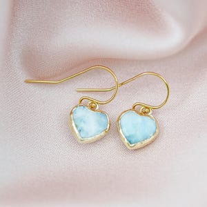 May include: A pair of heart-shaped earrings with a light blue stone and gold-colored trim. The earrings are displayed on a pink, silky fabric. The hooks are gold-colored.