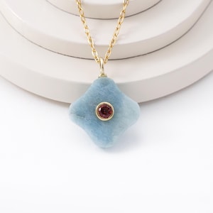May include: A gold chain necklace with a light blue, four-lobed pendant. The pendant has a small, round, red gemstone in the center. The necklace is displayed on a white, layered surface against a white background.
