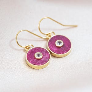 May include: A pair of gold-tone earrings with a circular, magenta-colored center and a small, clear gemstone in the middle. The earrings have a hook closure. The background is a soft, off-white fabric.