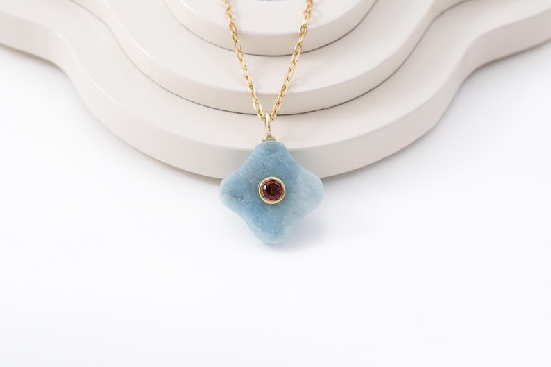 May include: A gold chain necklace with a light blue, four-lobed pendant. The pendant has a small, round, red gemstone in the center. The necklace is displayed on a white, layered surface against a white background.