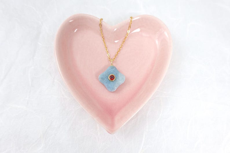 May include: A gold chain necklace with a light blue, four-petal pendant and a small red gemstone in the center. The necklace rests on a pink heart-shaped dish, set against a white background.