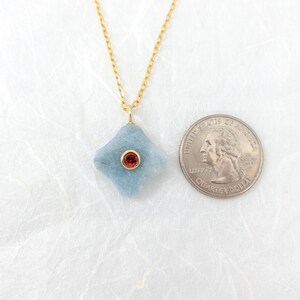 May include: A light blue pendant necklace with a gold chain. The pendant is a four-sided shape with a red gemstone in the center. A U.S. quarter is next to the pendant for size comparison.
