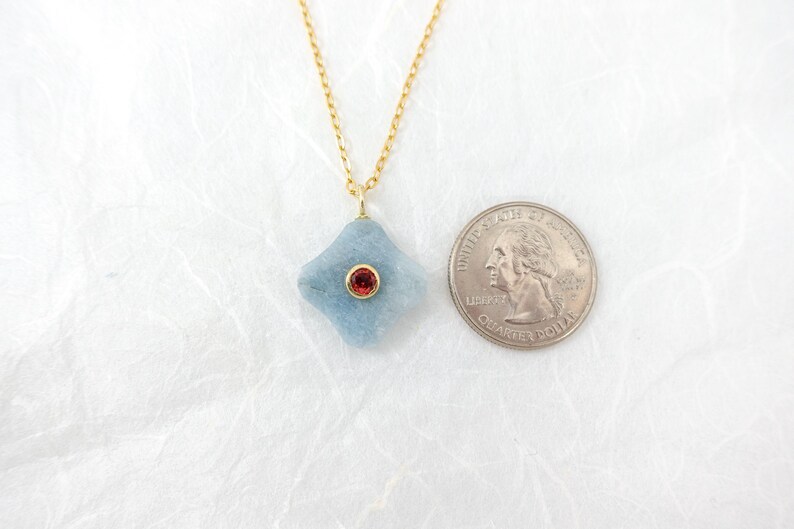May include: A light blue pendant necklace with a gold chain. The pendant is a four-sided shape with a red gemstone in the center. A U.S. quarter is next to the pendant for size comparison.