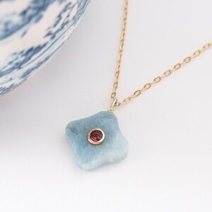 May include: A gold chain necklace with a light blue, irregularly shaped pendant. The pendant has a small, round, red gemstone set in the center. A blue and white patterned ceramic bowl is in the background.
