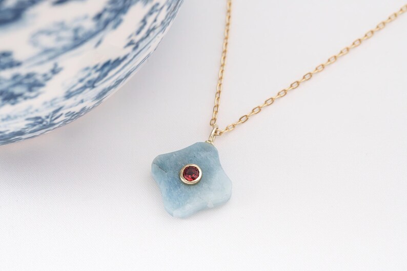 May include: A gold chain necklace with a light blue, irregularly shaped pendant. The pendant has a small, round, red gemstone set in the center. A blue and white patterned ceramic bowl is in the background.