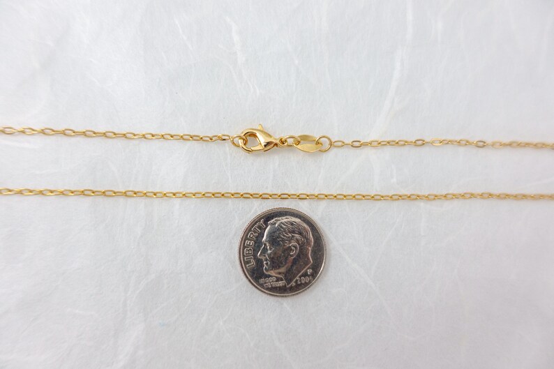 May include: A delicate gold-colored necklace with a lobster clasp, shown next to a dime for size comparison. The chain is thin and has small, evenly spaced links. The dime shows the profile of a man.