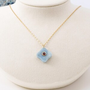 May include: A gold chain necklace featuring a four-pointed, light blue, matte-textured pendant. The center of the pendant is set with a small, round, dark red gemstone encased in a thin gold bezel, creating a contrast between the blue and red.
