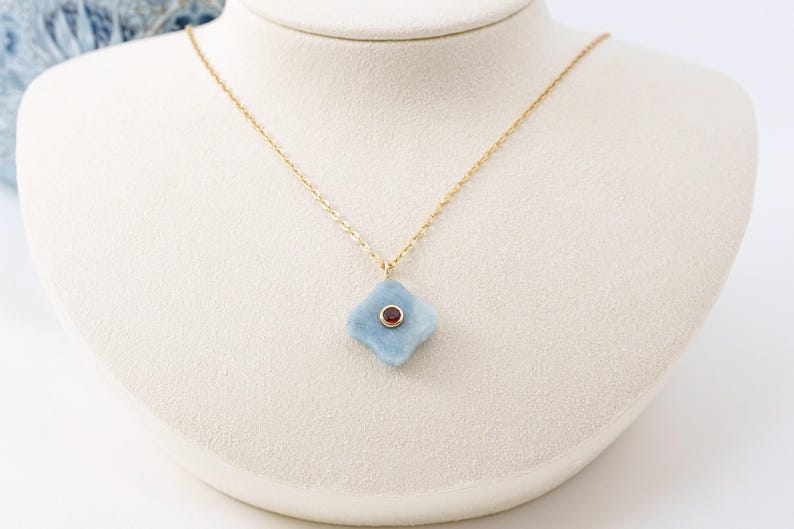 May include: A gold chain necklace featuring a four-pointed, light blue, matte-textured pendant. The center of the pendant is set with a small, round, dark red gemstone encased in a thin gold bezel, creating a contrast between the blue and red.