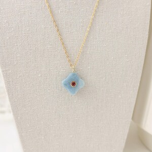May include: A delicate gold chain necklace featuring a light blue, four-petal flower pendant with a small, round, red gemstone center. The necklace is displayed on a neutral-colored fabric background.