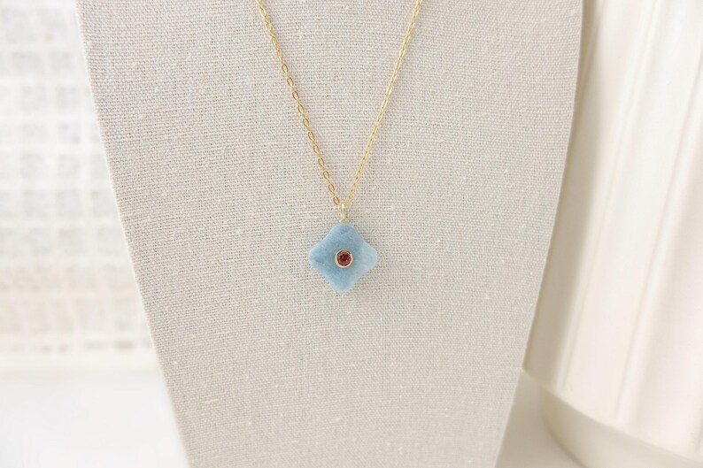 May include: A delicate gold chain necklace featuring a light blue, four-petal flower pendant with a small, round, red gemstone center. The necklace is displayed on a neutral-colored fabric background.