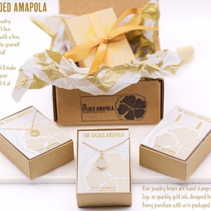 May include: A product shot of jewelry boxes from The Gilded Amapola. The boxes are kraft-colored with gold accents and a gold ribbon. The boxes contain necklaces and earrings. Text on the boxes reads "THE GILDED AMAPOLA".