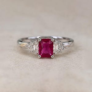 May include: A silver-toned ring featuring a central rectangular-cut vibrant red gemstone. The center stone is flanked by two smaller baguette-cut clear stones and a cluster of tiny round clear stones on each side, set on a polished metal band.