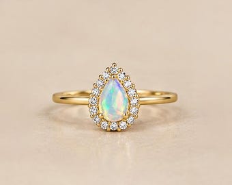 May include: A gold ring featuring a pear-shaped opal center stone surrounded by a halo of small round white diamonds. The band is thin and polished gold, creating a delicate look with the iridescent gemstone and sparkling accent stones.
