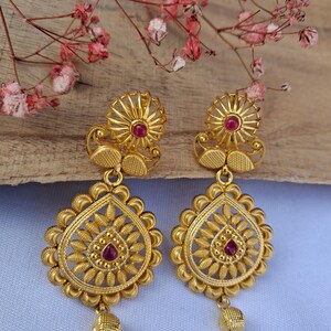 Könnte beinhalten: Ein Paar goldfarbene Ohrringe mit floralem Design. Jeder Ohrring hat einen kleinen, runden, roten Edelstein in der Mitte der oberen und unteren floralen Elemente. Die Ohrringe haben eine Tropfenform und eine kleine, strukturierte Kugel am unteren Ende.