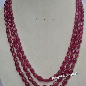 May include: A three-strand necklace featuring polished, oval-shaped deep pink ruby beads. The strands are arranged in a cascading layered design. The text "Priyanka creation" is printed in white on the lower right side of the necklace strands.