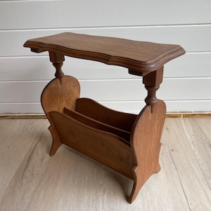 Peut inclure: Une table d'appoint en bois brun dotée d'un plateau rectangulaire aux bords incurvés, soutenue par deux piliers tournés qui se connectent à une section porte-revues inférieure avec des panneaux latéraux incurvés et un séparateur central.