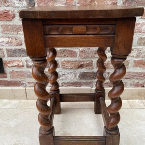 May include: A small, antique wooden side table with a dark brown finish. The table features a rectangular top, a decorative carved apron, and four spiral-turned legs. The legs are connected by a lower stretcher for added stability. The table is set against a brick wall.