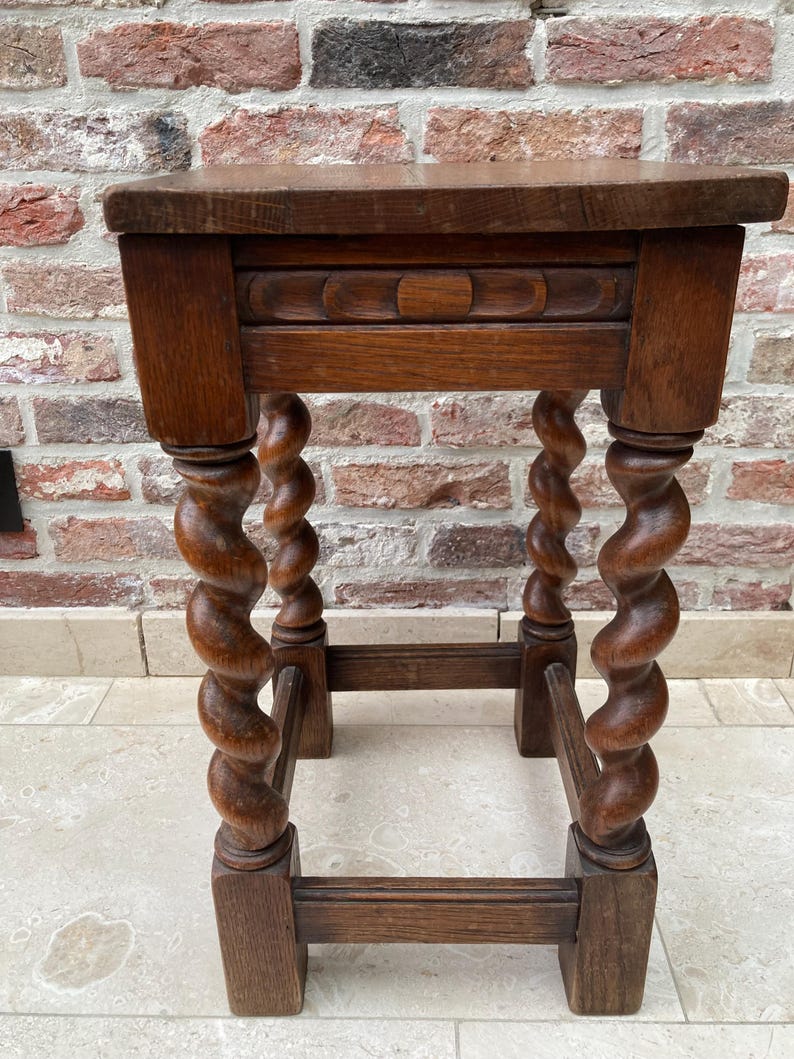 May include: A small, antique wooden side table with a dark brown finish. The table features a rectangular top, a decorative carved apron, and four spiral-turned legs. The legs are connected by a lower stretcher for added stability. The table is set against a brick wall.