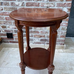 May include: A round, dark brown wooden side table with a decorative floral inlay on the tabletop. The table has a lower shelf and four turned legs. The table is approximately 61 cm in diameter and 71 cm tall.