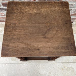 May include: A small, square wooden side table with a dark brown finish. The tabletop shows visible wood grain and some light markings. The legs feature a decorative, turned design. The table is set against a brick wall and tiled floor.