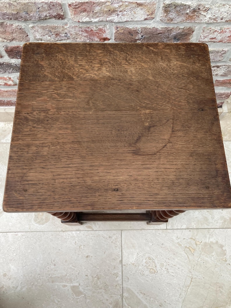 May include: A small, square wooden side table with a dark brown finish. The tabletop shows visible wood grain and some light markings. The legs feature a decorative, turned design. The table is set against a brick wall and tiled floor.