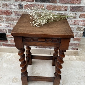 May include: A small, antique wooden side table with a square top and intricately carved, spiral-turned legs. The table is a warm brown color and has a decorative carved apron. A small bouquet of white flowers rests on the tabletop.