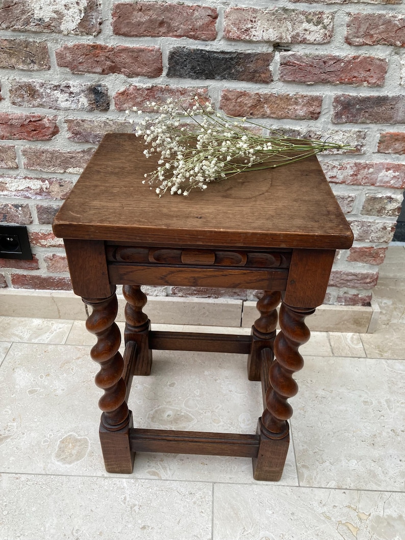 May include: A small, antique wooden side table with a square top and intricately carved, spiral-turned legs. The table is a warm brown color and has a decorative carved apron. A small bouquet of white flowers rests on the tabletop.